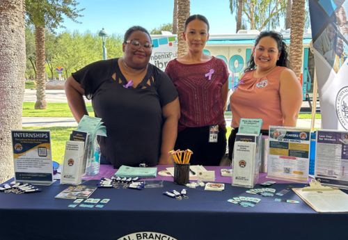 Three women standing behind a table, smiling and wearing purple ribbons, standing outdoors with informational materials