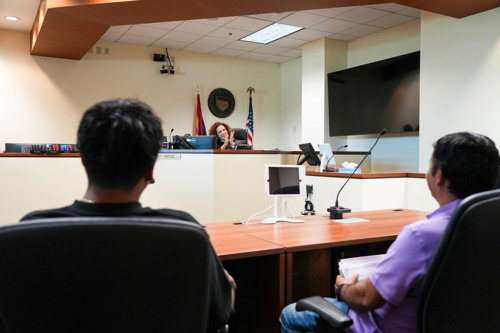 Two youth are seated behind a table facing Judge Hartley's bench as she smiles and claps her hands to address the youth from the bench in her courtroom.