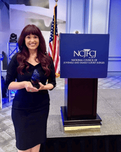 Picture of Judge Lori Bustamante posing with a blue glass award next to a podium for NCJFCJ National Council of Juvenile and Family Court Judges